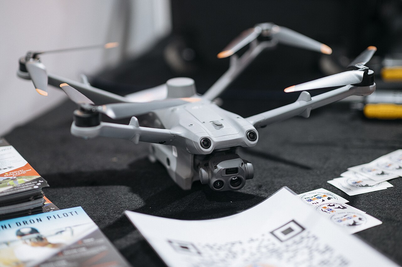 A silver camera drone sitting on a table with flyers and papers at an indoor technology event.