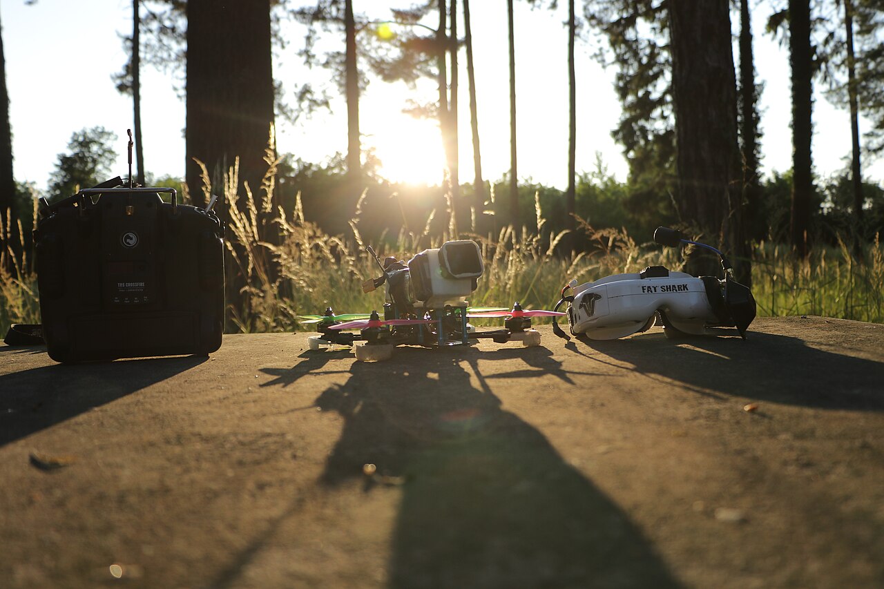An FPV radio, racing-style quadcopter, and Fat Shark goggles on a concrete slab at sunset, with trees behind them.